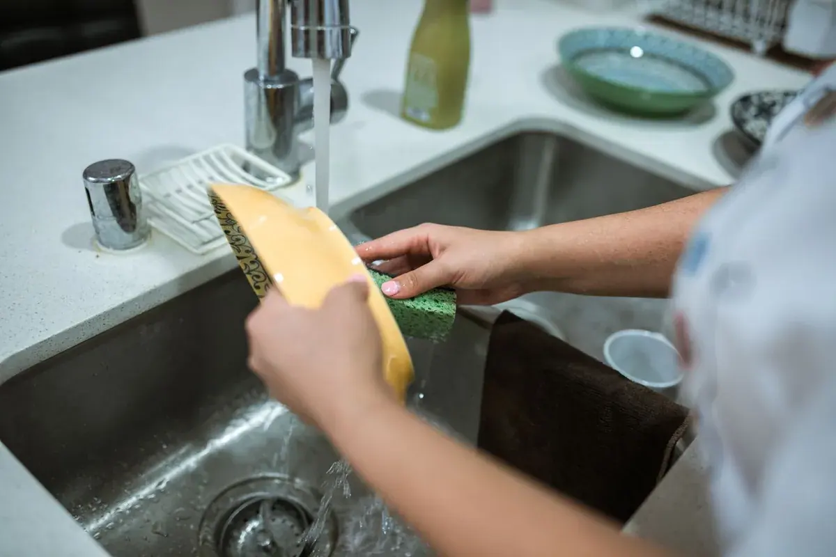 Mãos lavando o cesto da air fryer na pia com esponja e água corrente